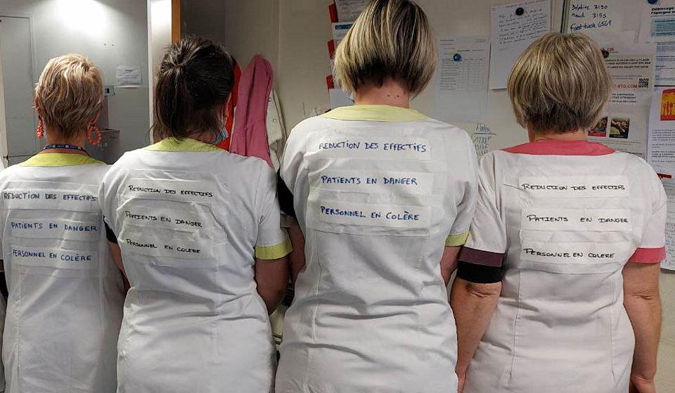 Quatre femmes en blouse blanches portent sur leur dos le messages "réduction des effetifs", "patients en danger", "personnel en colère"